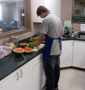 Youth in a blue apron cutting up large watermelon 